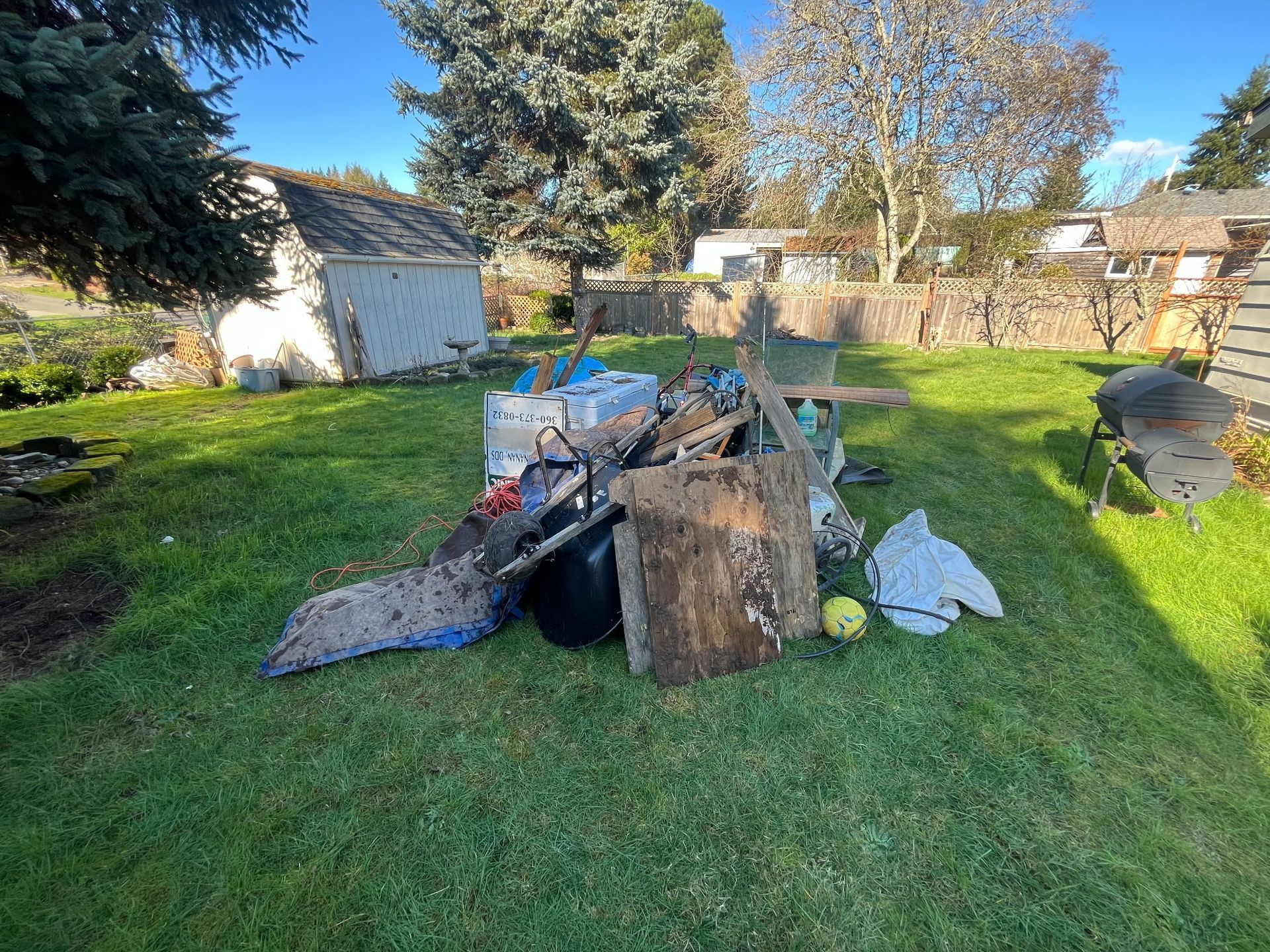 Backyard pile of yard and household waste, wood planks, tarps, and a wheelbarrow on green grass