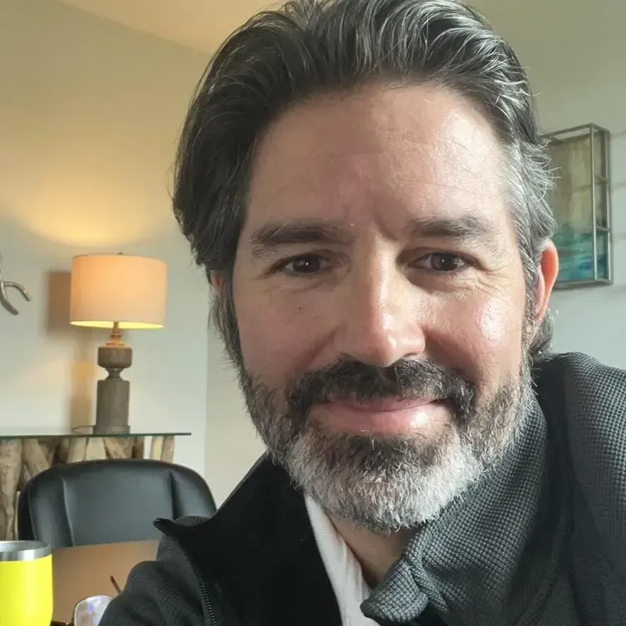 Smiling man with gray beard in a cozy living room, with a lamp and chair behind him