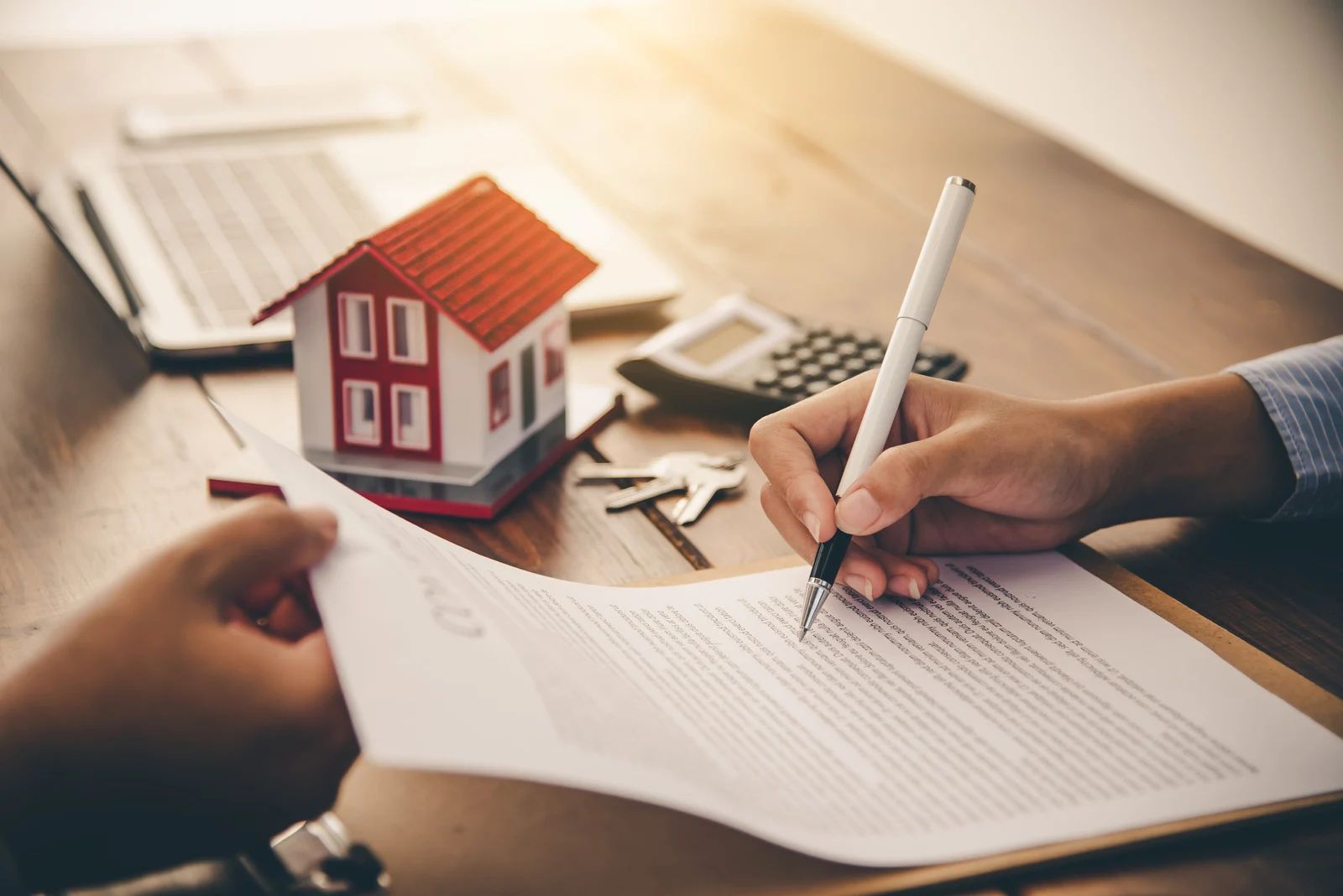 Hands signing a real estate contract beside a house model and keys on a desk