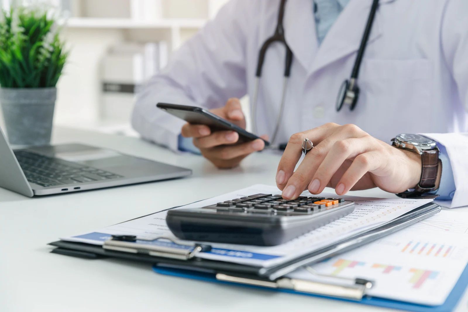 Doctor using a calculator and smartphone at a desk with laptop, clipboard, and papers