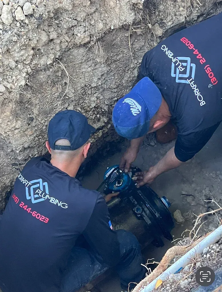 Two plumbers in black shirts with company logos work on a pipe in a trench.