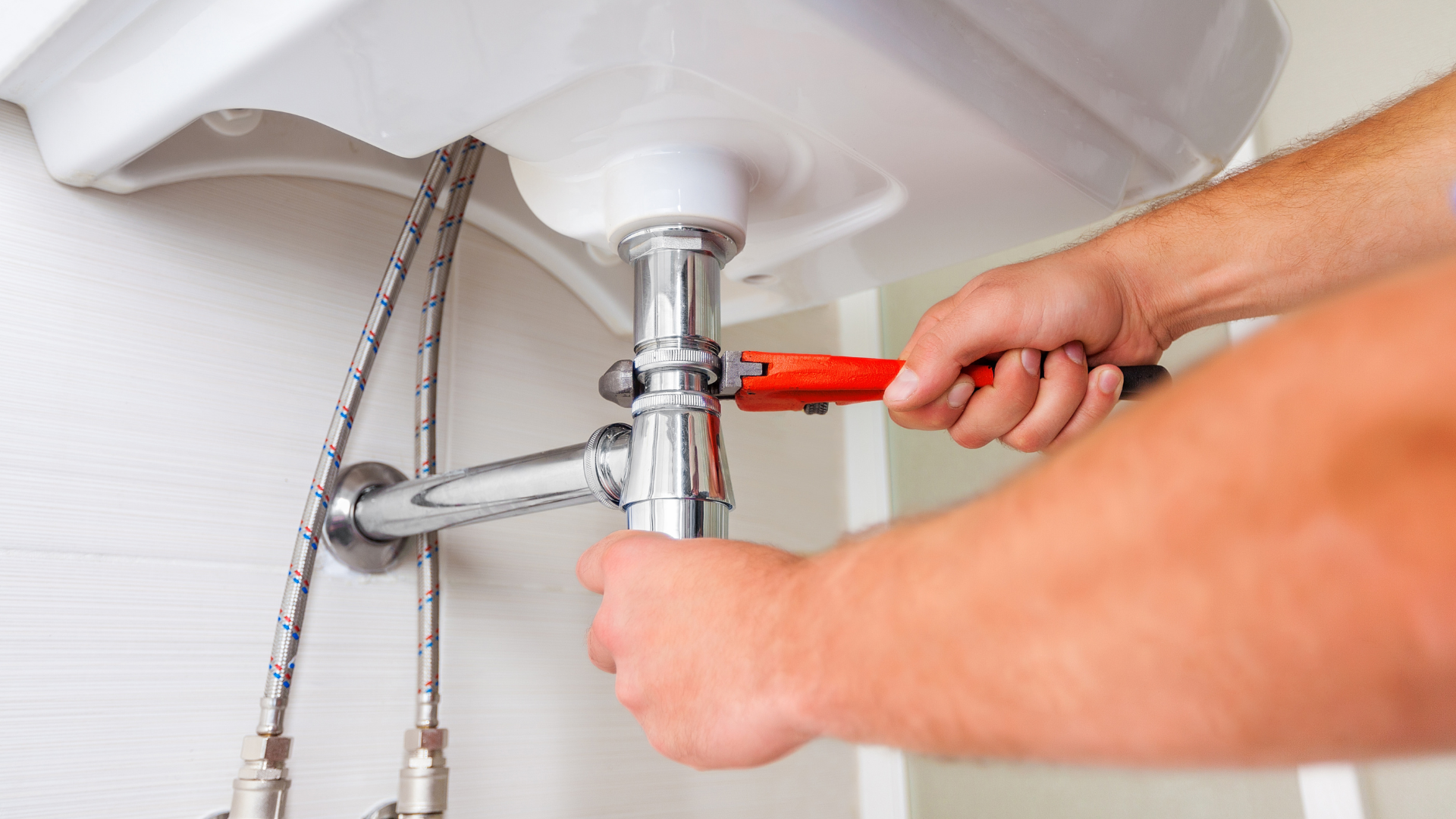Plumber using a wrench under a white sink.  He is tightening a pipe fitting, a chrome-colored drainpipe.