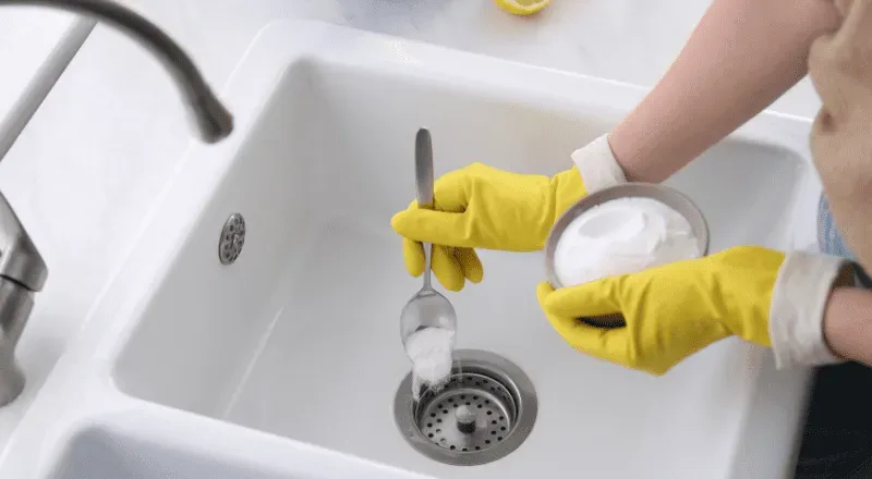 Person wearing yellow gloves pouring baking soda into a white sink drain with a spoon.