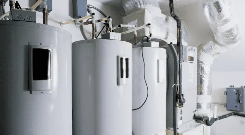 Several white water heaters in an indoor utility room, with various pipes and ductwork visible.