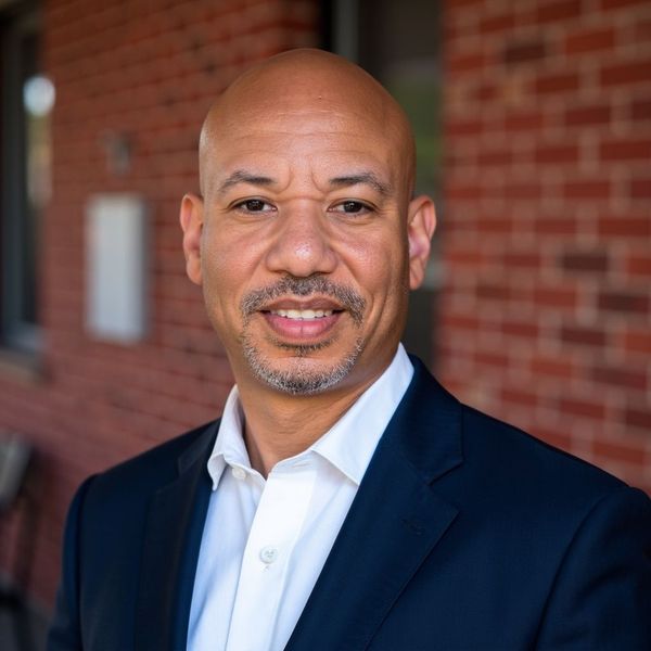 Smiling man in a navy blazer and white shirt standing outdoors by a brick wall