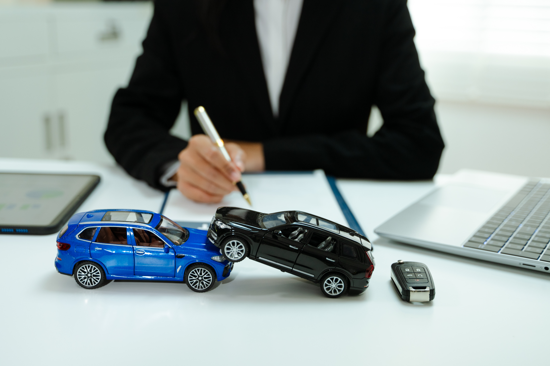 Miniature car accident scene on a desk with a person writing, blue and black toy cars, and laptop nearby
