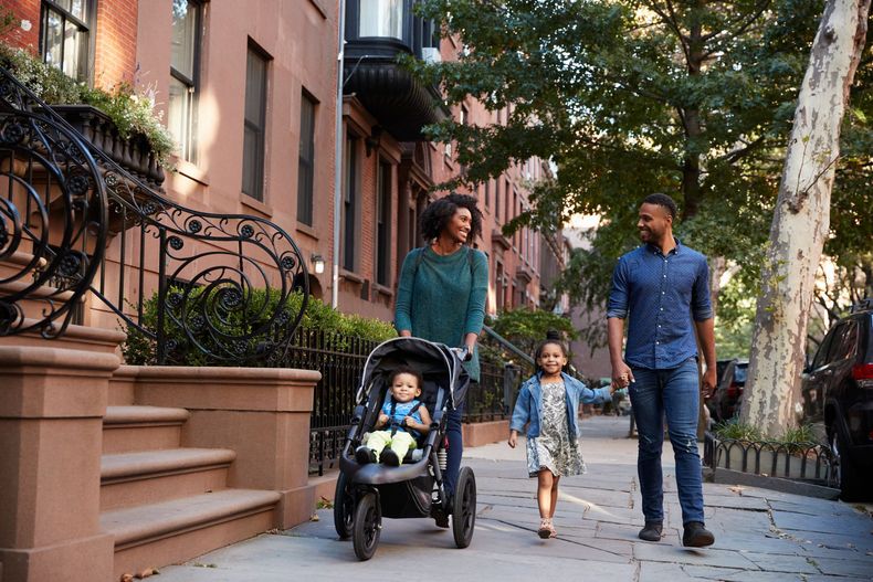 Family walking on a tree-lined city sidewalk, with a child in a stroller and apartment buildings beside them