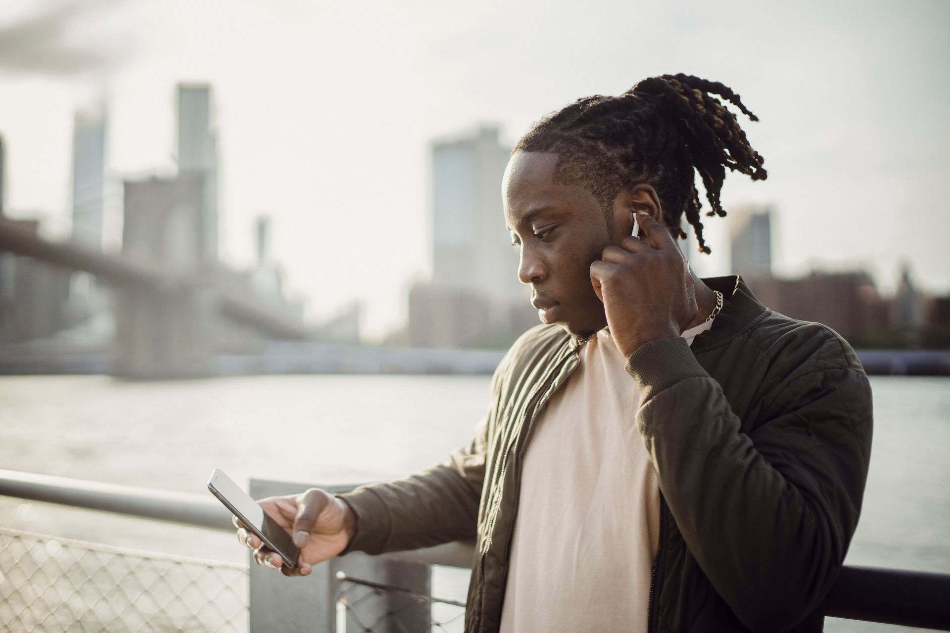 Person on a riverside walkway looking at a phone, with a bridge and city skyline in the background.
