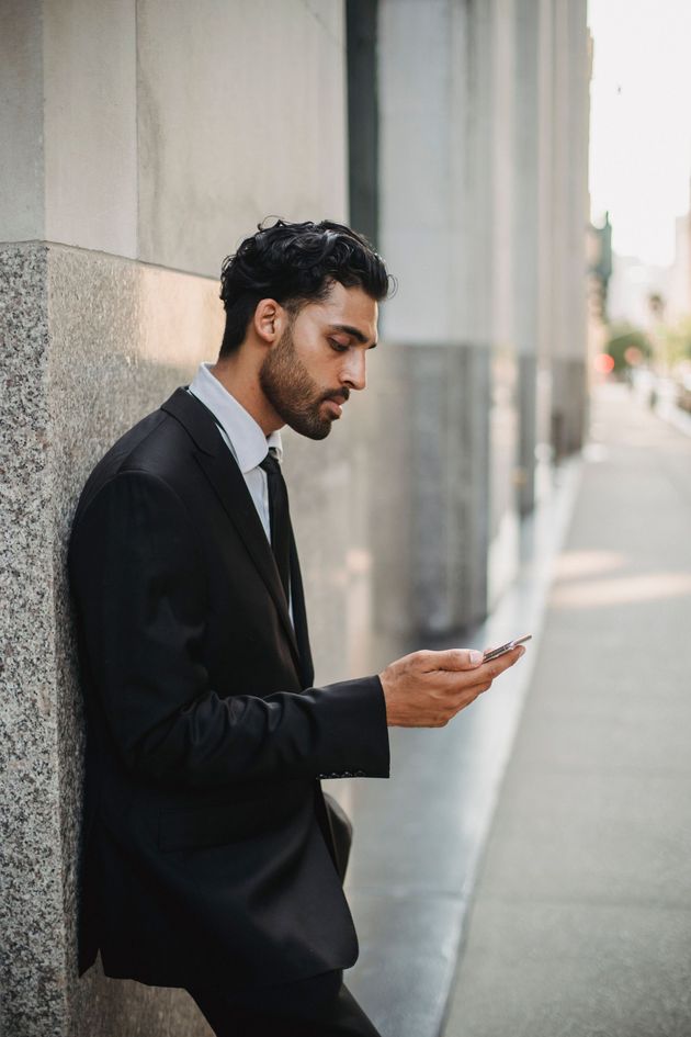 Man in a black suit leaning against a wall, looking at a smartphone outdoors.