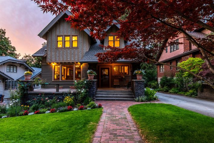 Two-story house with warm lit windows, brick path, green lawn, and red tree at dusk