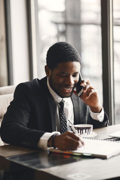 Man in a suit smiling while talking on a phone and writing at a desk by a window
