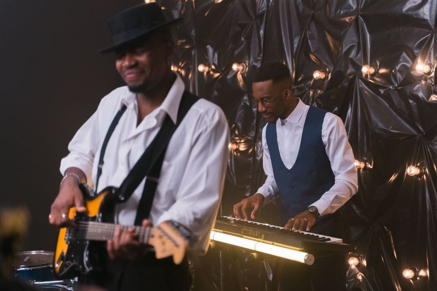 Two musicians perform on stage, one playing guitar and one behind a keyboard, in dim warm lighting.