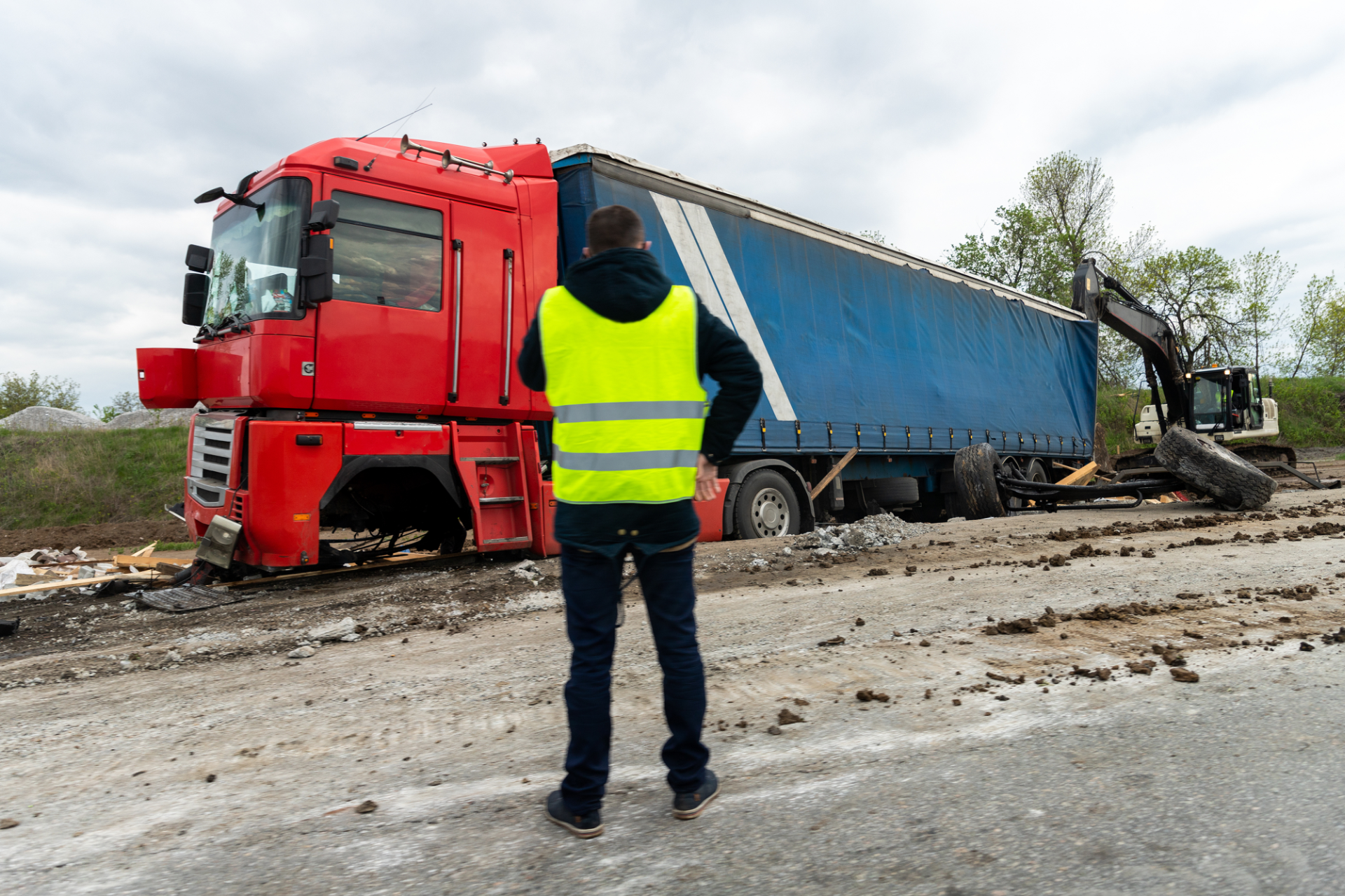 Worker in neon vest photographing a red truck and blue trailer stuck in muddy roadside debris.