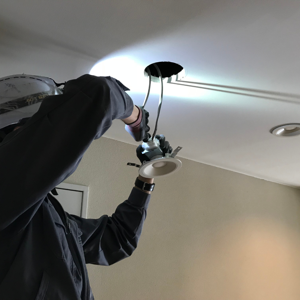 Person installing a recessed light in a ceiling; holding the light fixture, wires visible, bright spotlight.