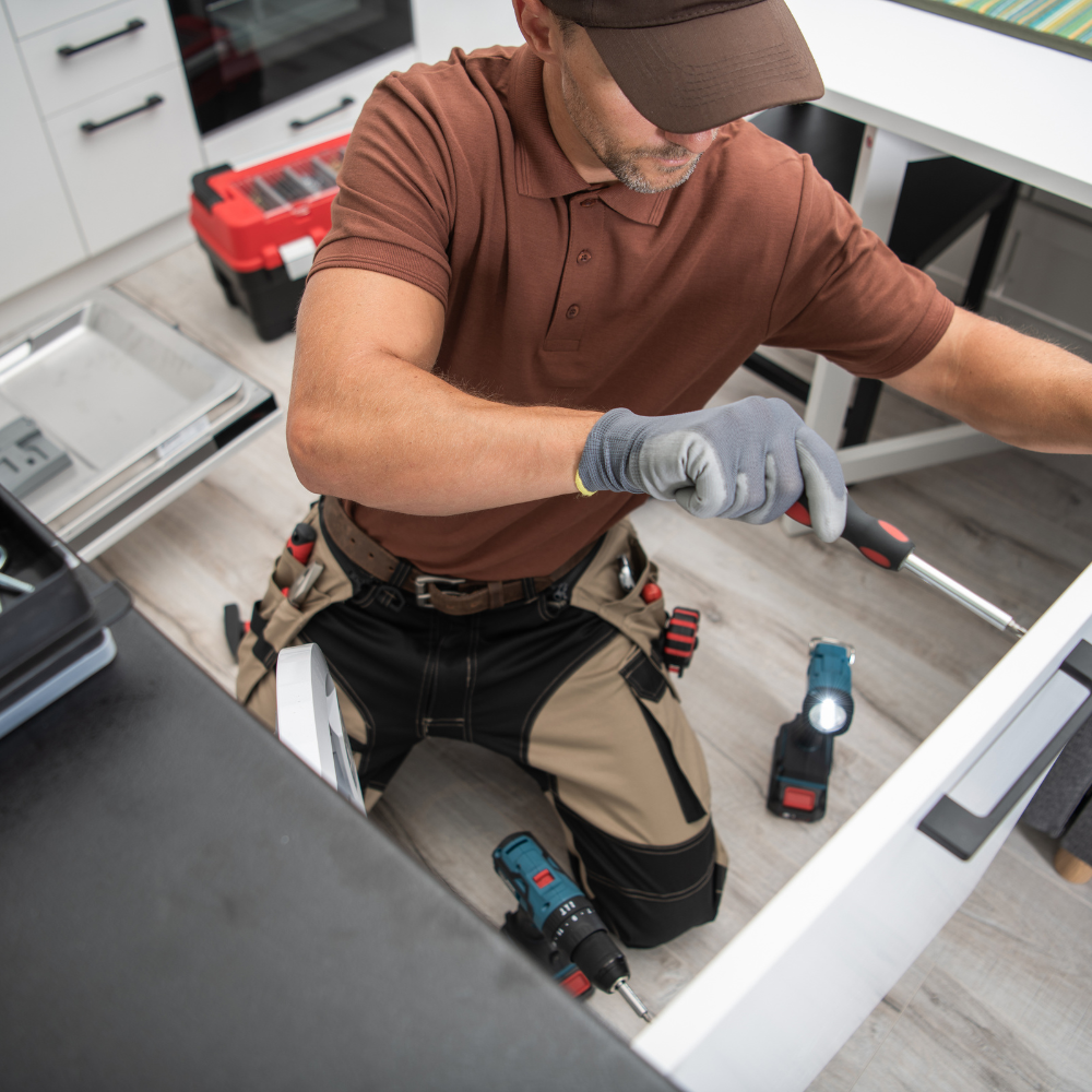 Man in brown shirt and cap installing cabinet, using a screwdriver, kneeling in a kitchen.