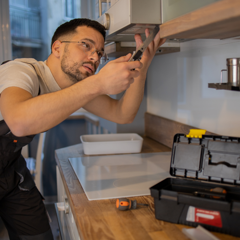 A person in overalls using a screwdriver on a kitchen range hood. Toolbox on the counter.