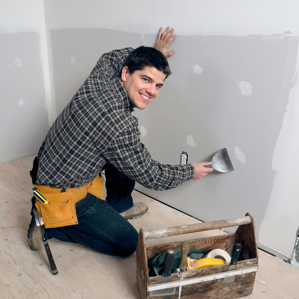 Man using a putty knife to repair drywall in a room with a tool box.