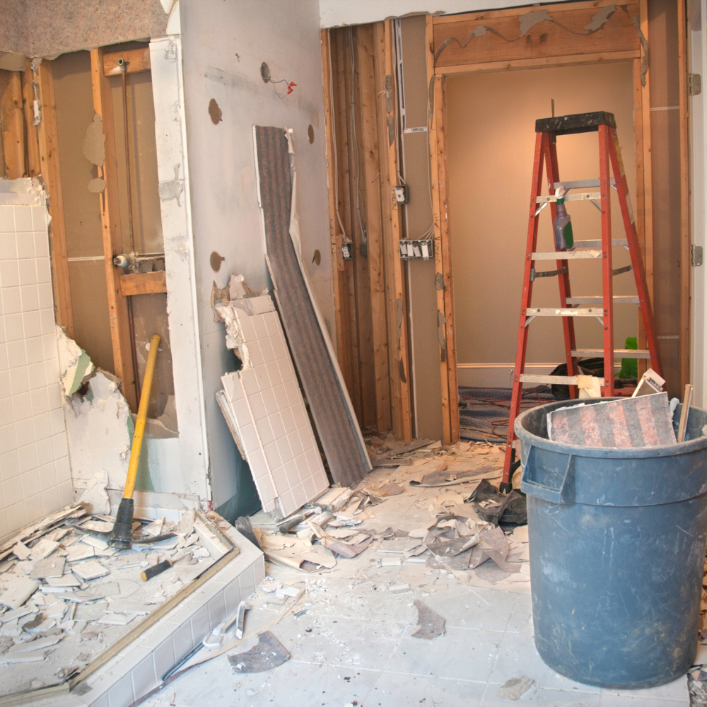 Bathroom under renovation: walls torn down, debris on floor, ladder, trash bin.