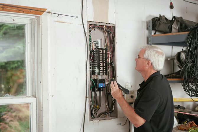 Man examining an open electrical panel with a flashlight in a garage.