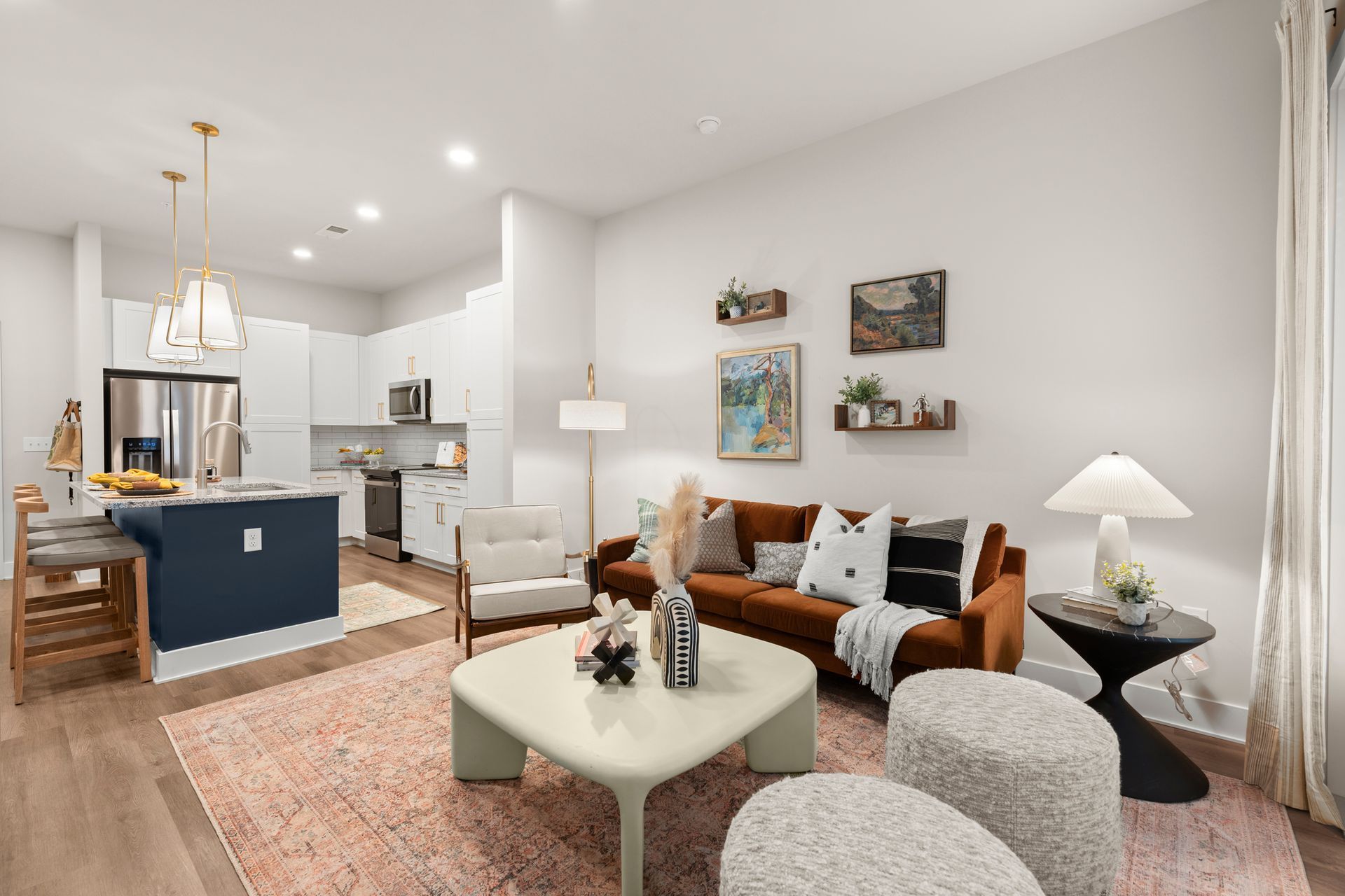 Living room with rust-colored sofa, white chairs, and a kitchen with navy island; rug.