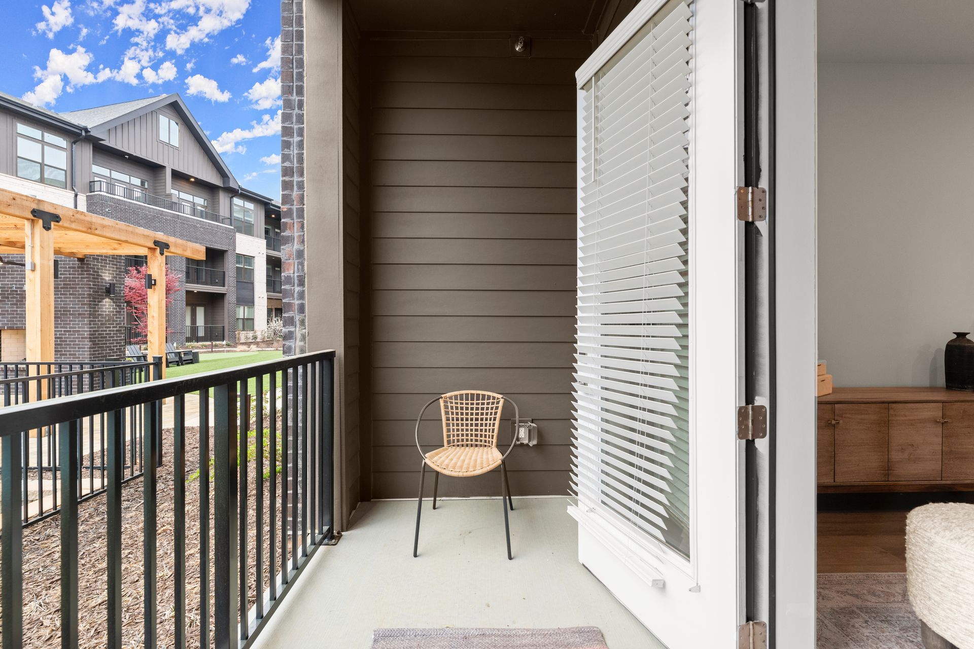 Balcony with wicker chair, open door. Brown siding, black railing, other buildings and blue sky visible.