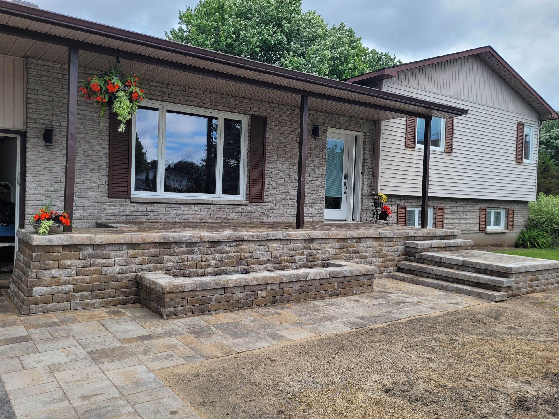 Brick house with stone patio, porch, and steps. Hanging flowers by a window, a front door, and the neighboring house.