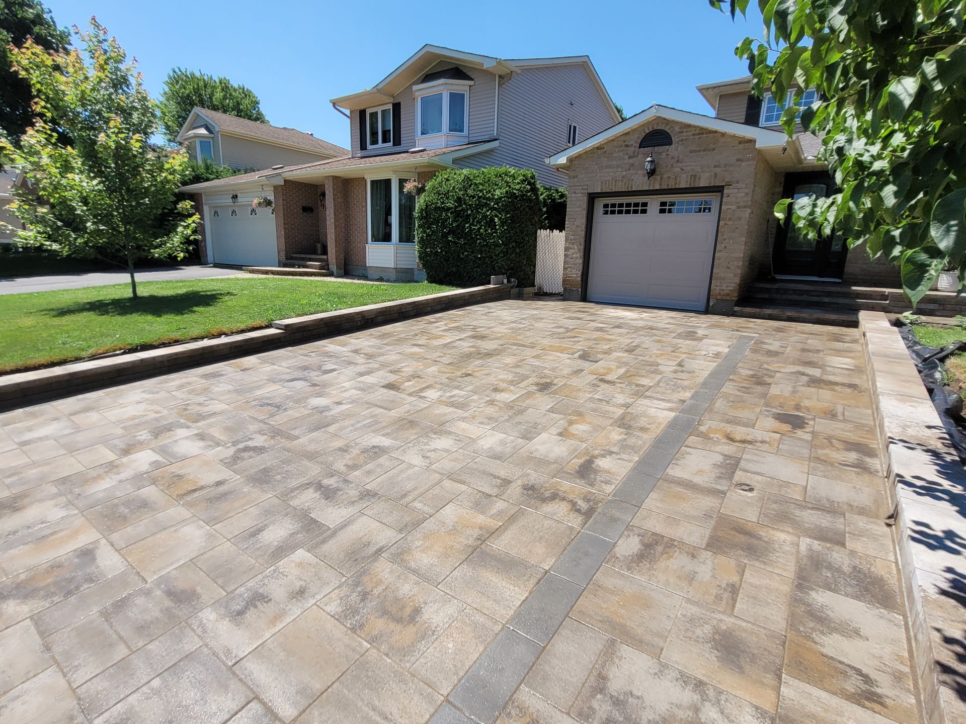 Paver driveway leading to a beige house with garage. Sunny day, green lawn, and blue sky.