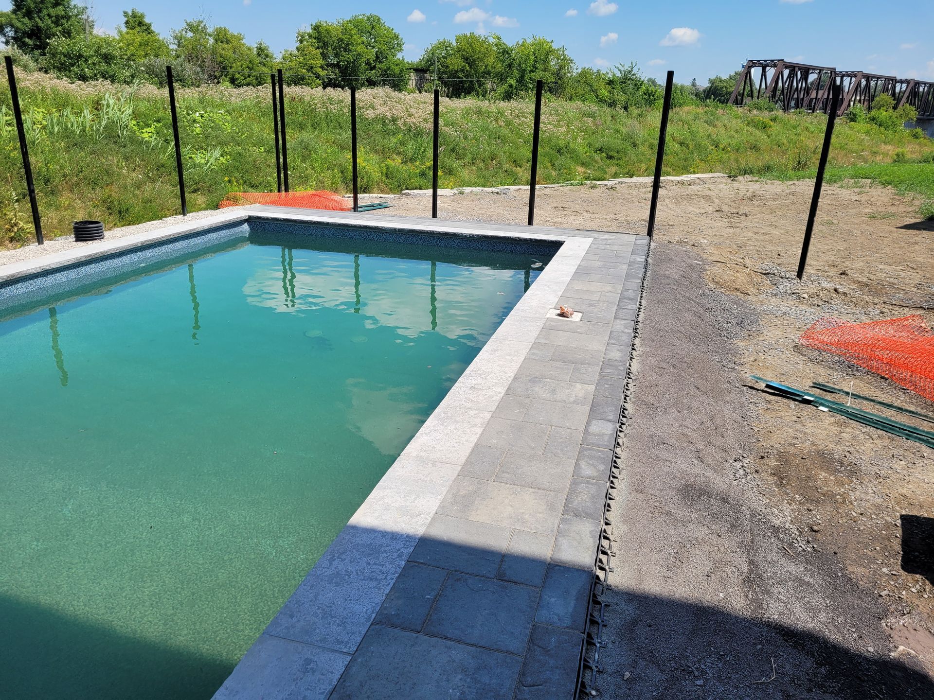 A rectangular pool with light green water, surrounded by pavers, gravel, and black fence posts, set in a grassy field.