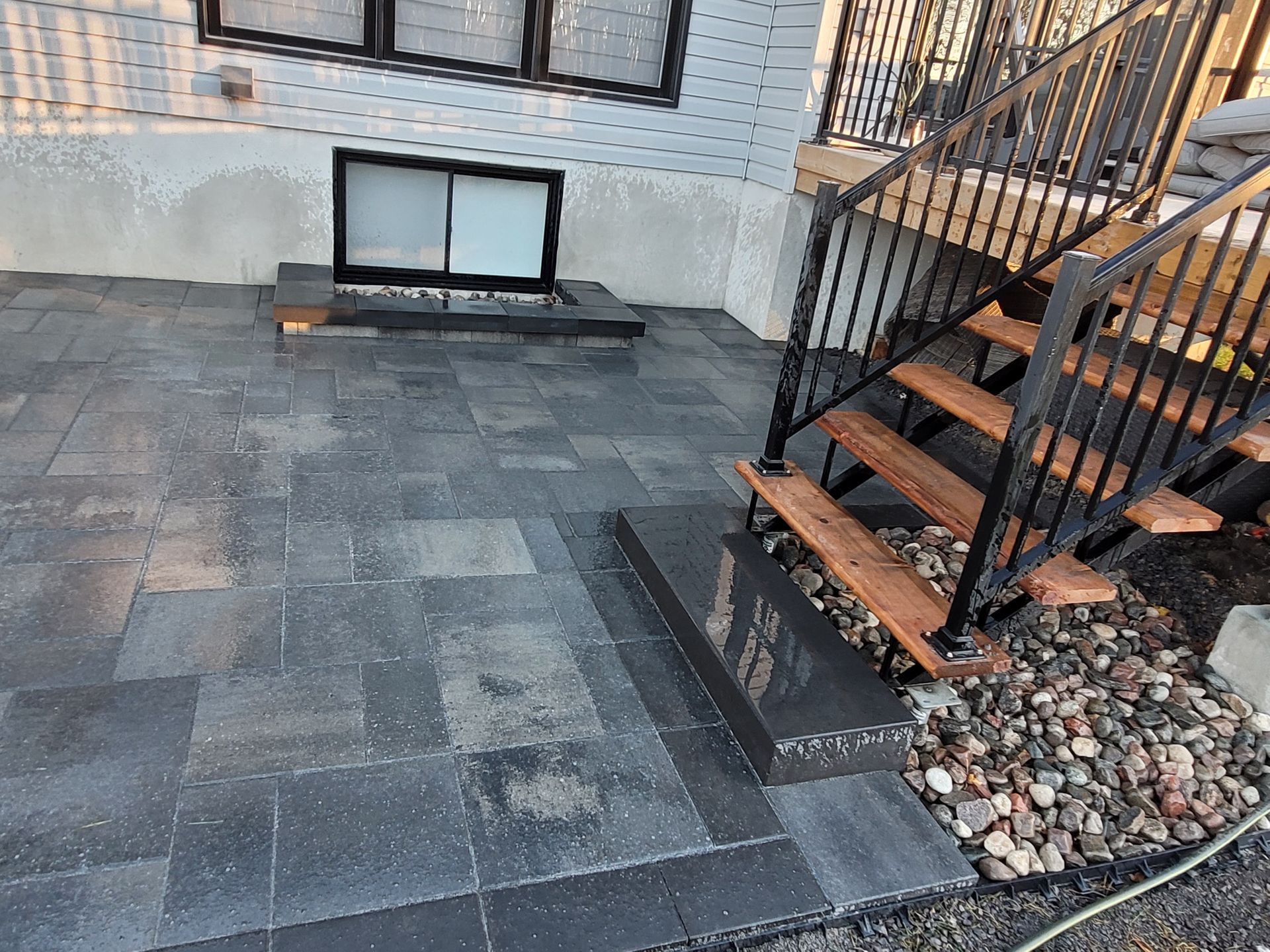 Gray paver patio with steps, leading to a black metal staircase; window well in background.