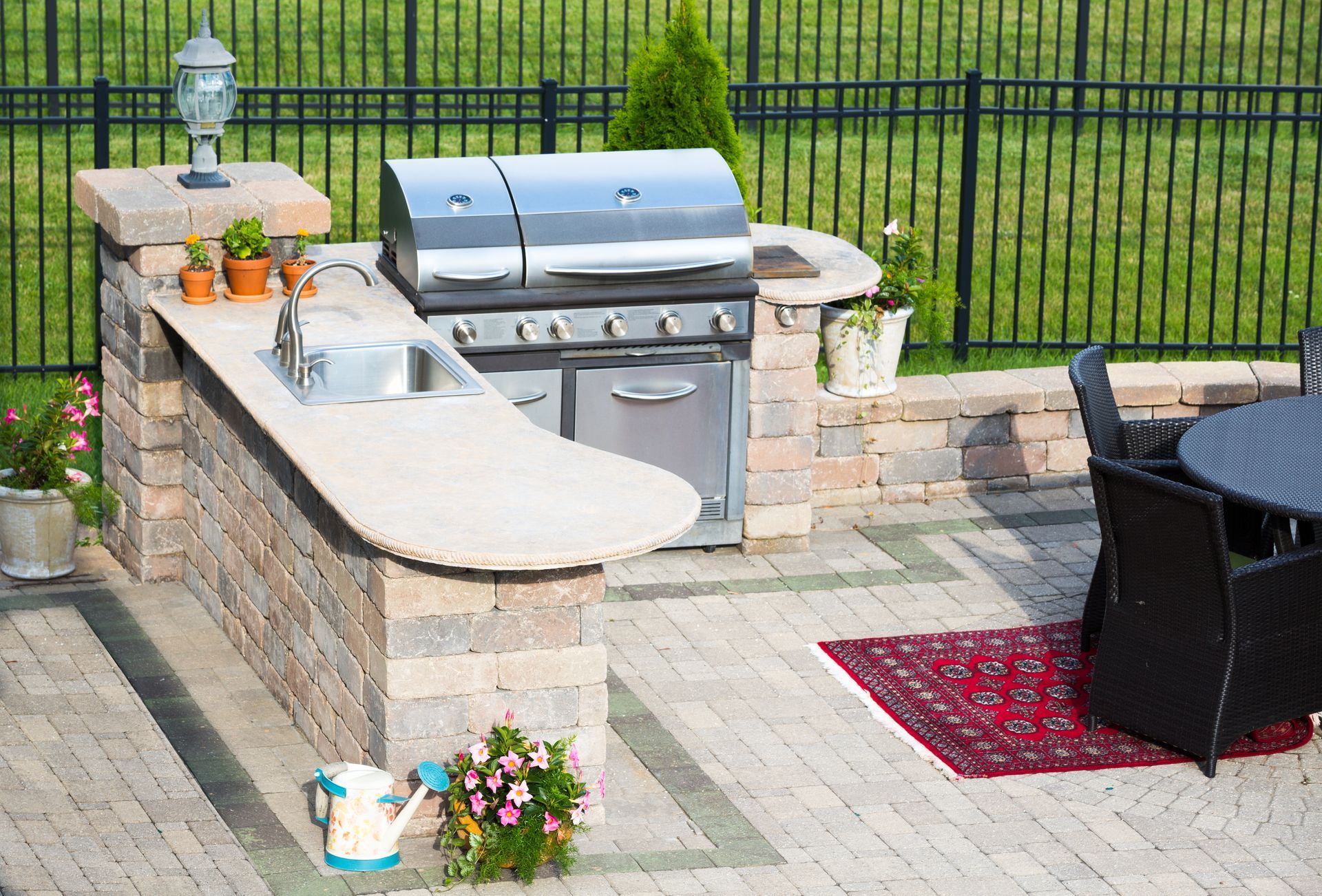 Outdoor kitchen with grill, sink, and countertop. Dining table, fence, and plants.