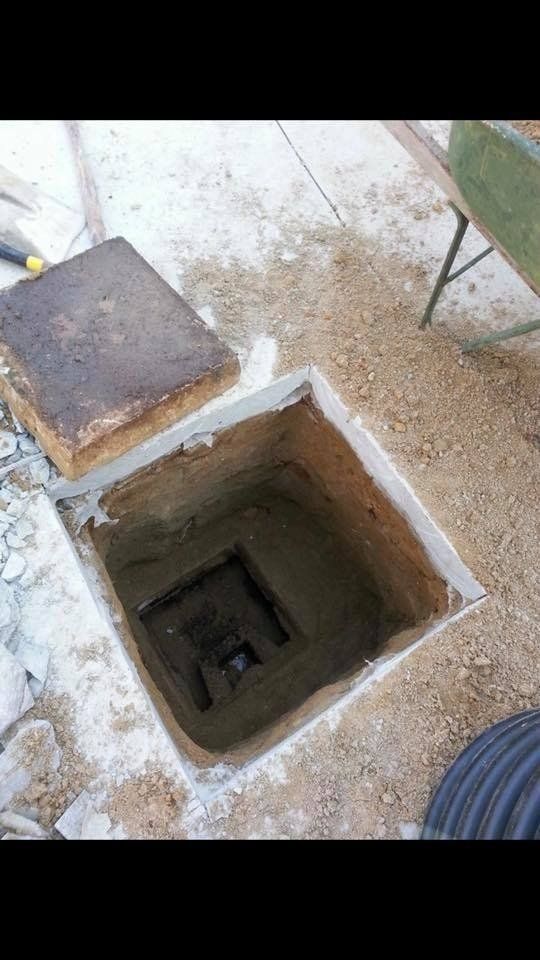 Open concrete access point with lid, surrounded by gravel and a wheelbarrow.