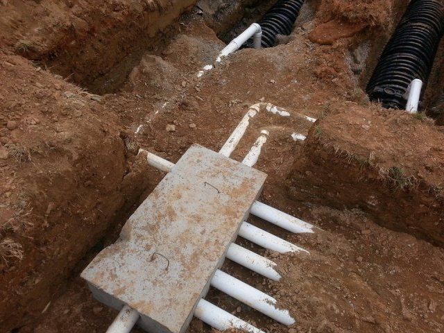 Excavated trench with white and black pipes, a concrete block, and brown soil.