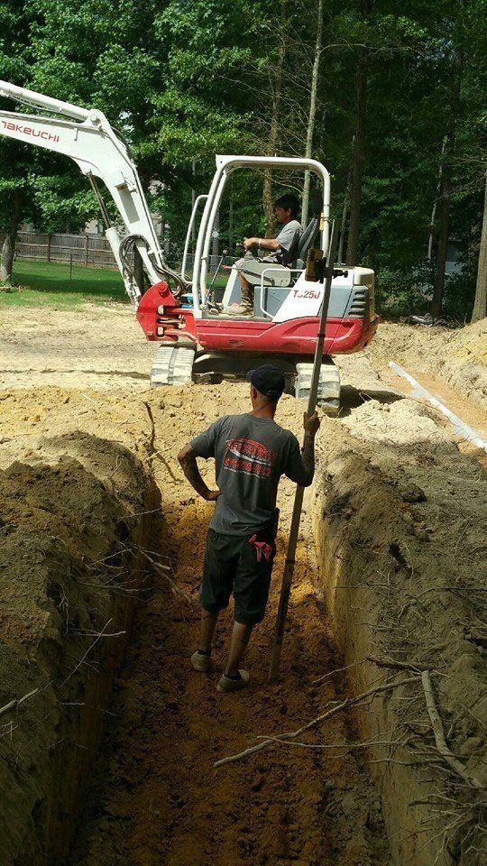 Man in a trench, using a stick while an excavator operator is in the background. Construction site, sunny day.