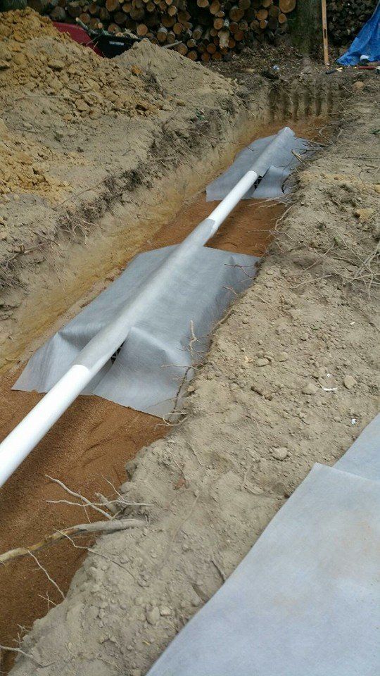 White PVC pipe in a trench wrapped with gray fabric, soil, and concrete slabs.