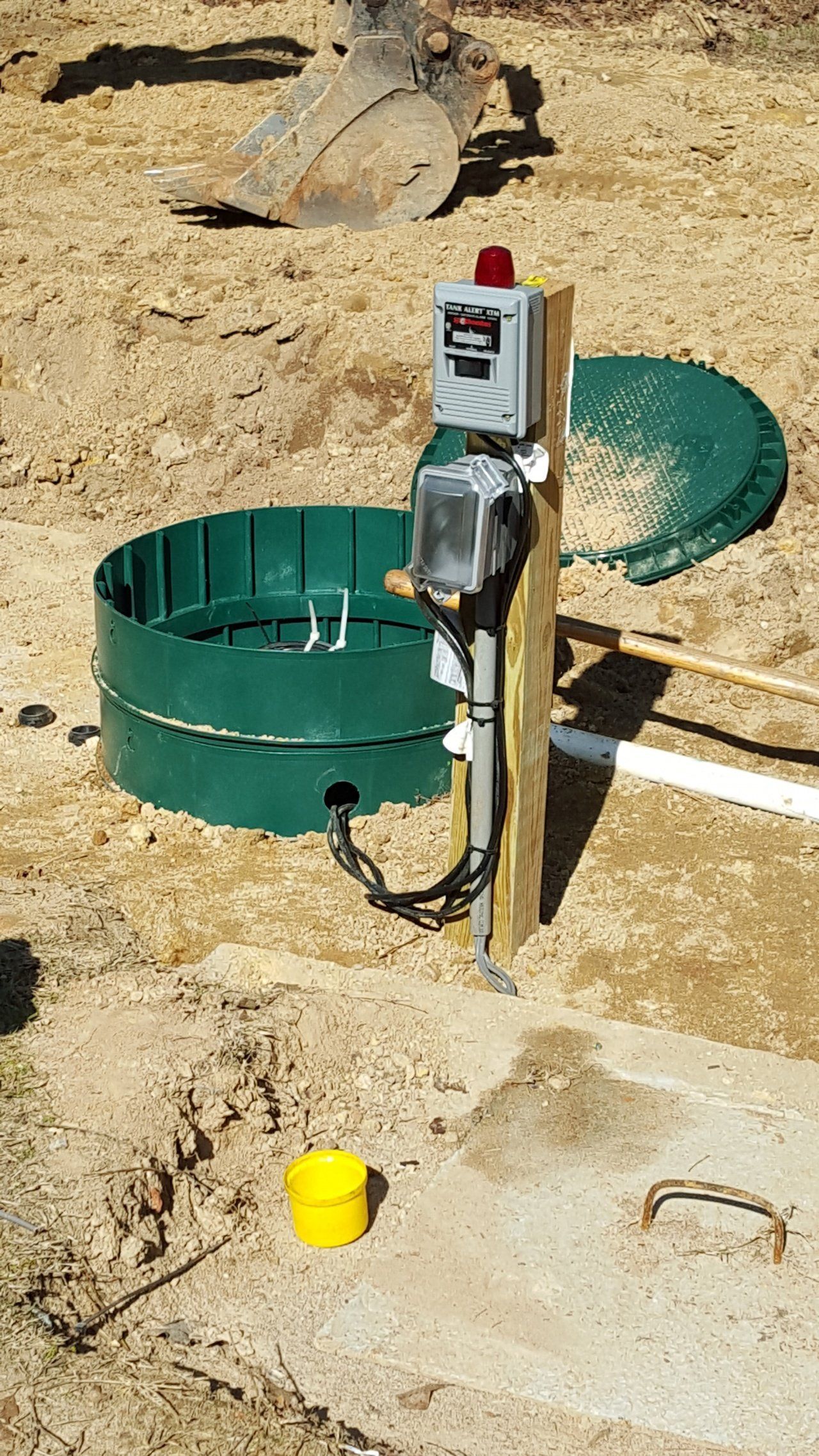 Septic system components: green tank, control box on post, warning light, and excavator in dirt.