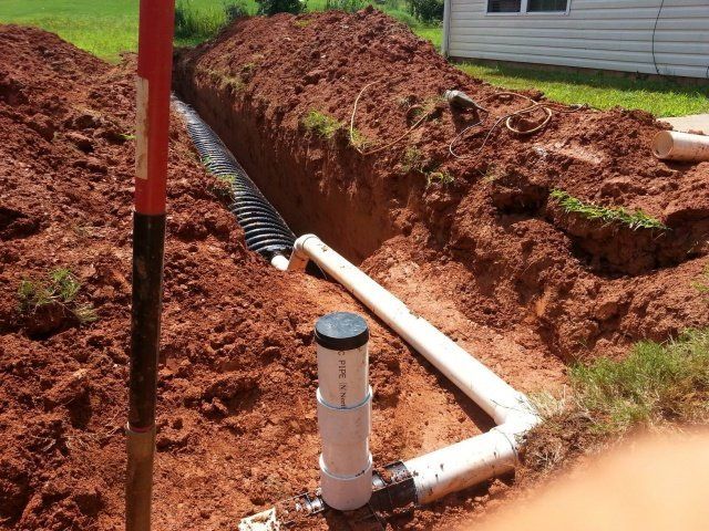 Trench with drain pipe, white PVC pipes, and soil. Installation outside near a house.
