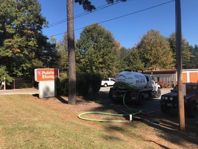 A septic service truck connected to a drain near a Public Storage sign and trees on a sunny day.