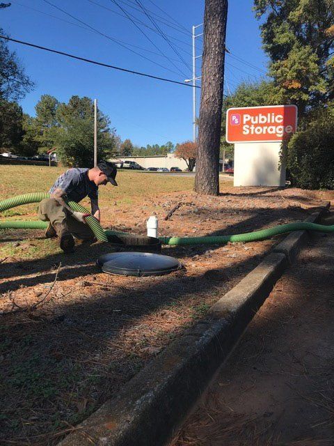 Man kneeling, connecting a green hose to a septic tank near a Public Storage sign. Outdoors, sunny day.