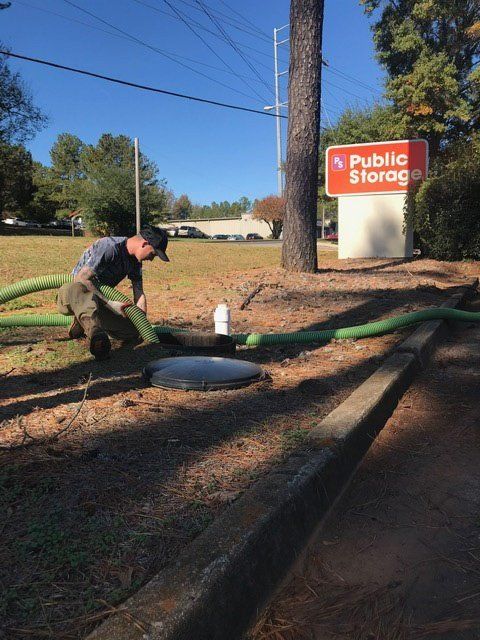 Man connects hose to a septic tank access point near Public Storage sign. Sunny day.