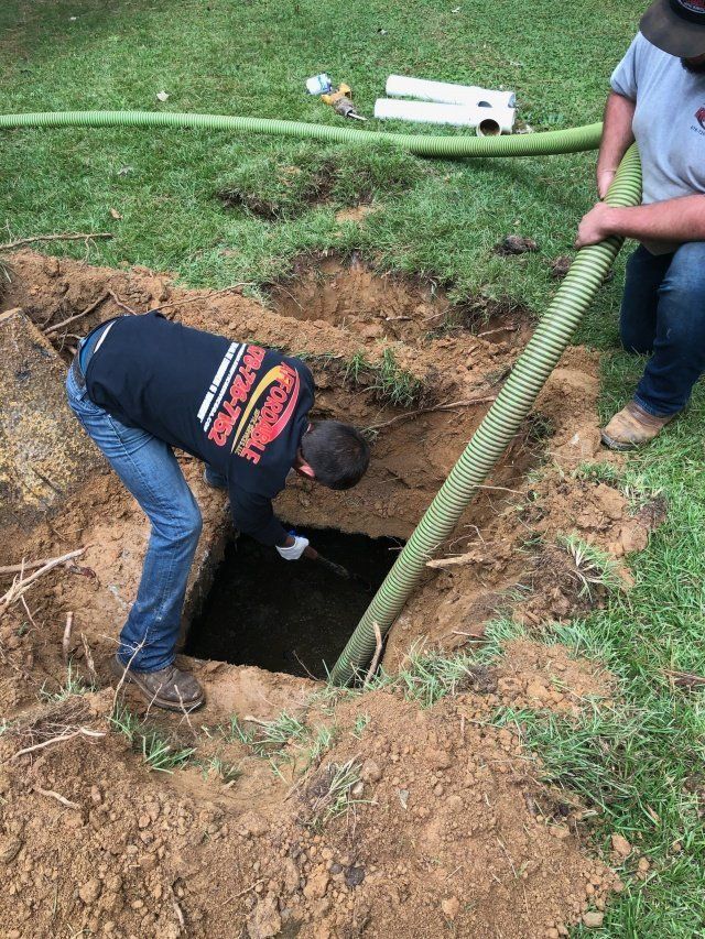 Two people working at a square hole in a lawn, one using a hose. The hole contains dark liquid.