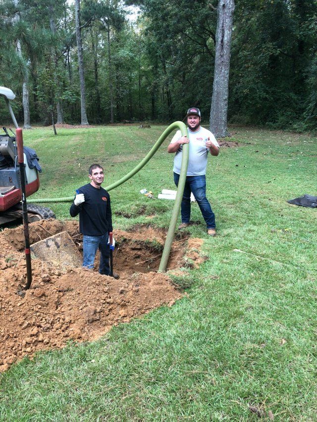 Two men with a hose near a rectangular hole in a grassy yard. One is inside the hole, the other holds the hose.