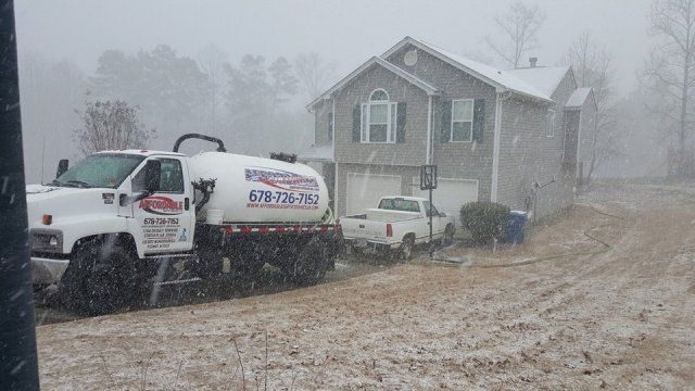 A white tanker truck parked in front of a gray house in a snowstorm.