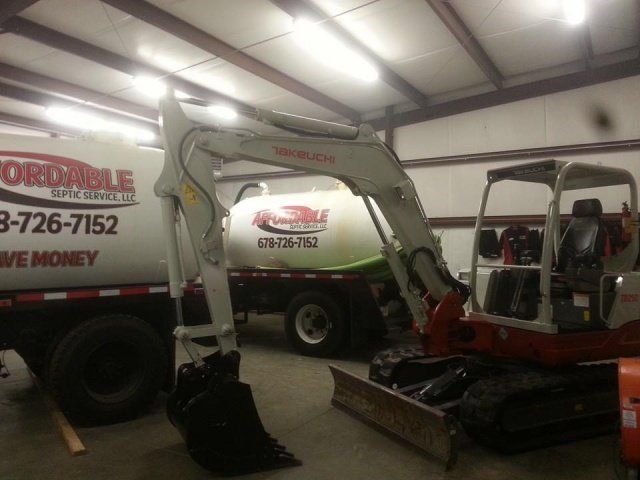 Mini excavator with bucket and blade, parked inside a garage. Affordable Septic truck in background.