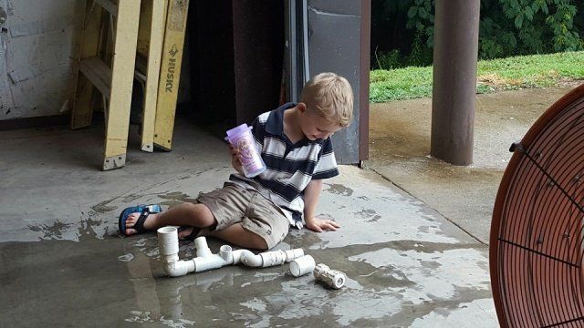Boy with blond hair, striped shirt, and shorts sits on wet concrete floor, looking at white PVC pipes. A fan and ladder are nearby.