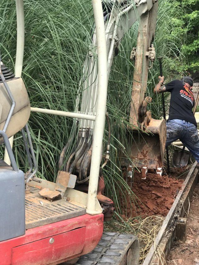 An excavator digs a trench next to tall grasses, a man operates the machine.