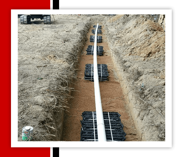 Trench with gravel and black plastic containers, connected by a white pipe. A backhoe is in the background.