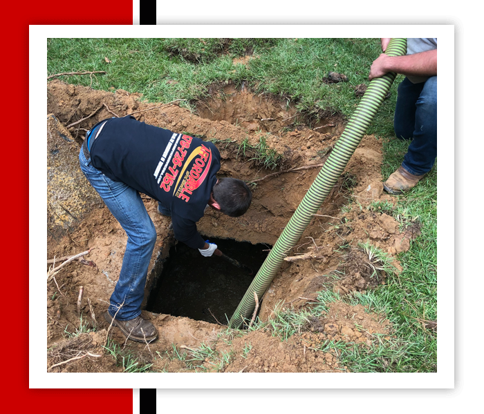 Man cleaning a septic tank in a muddy hole. A hose extends into the dark water. Someone else holds the hose.