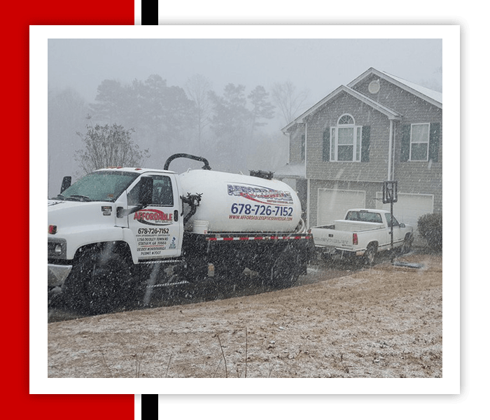 Septic tank truck in snowy residential area with a house and a pickup truck.