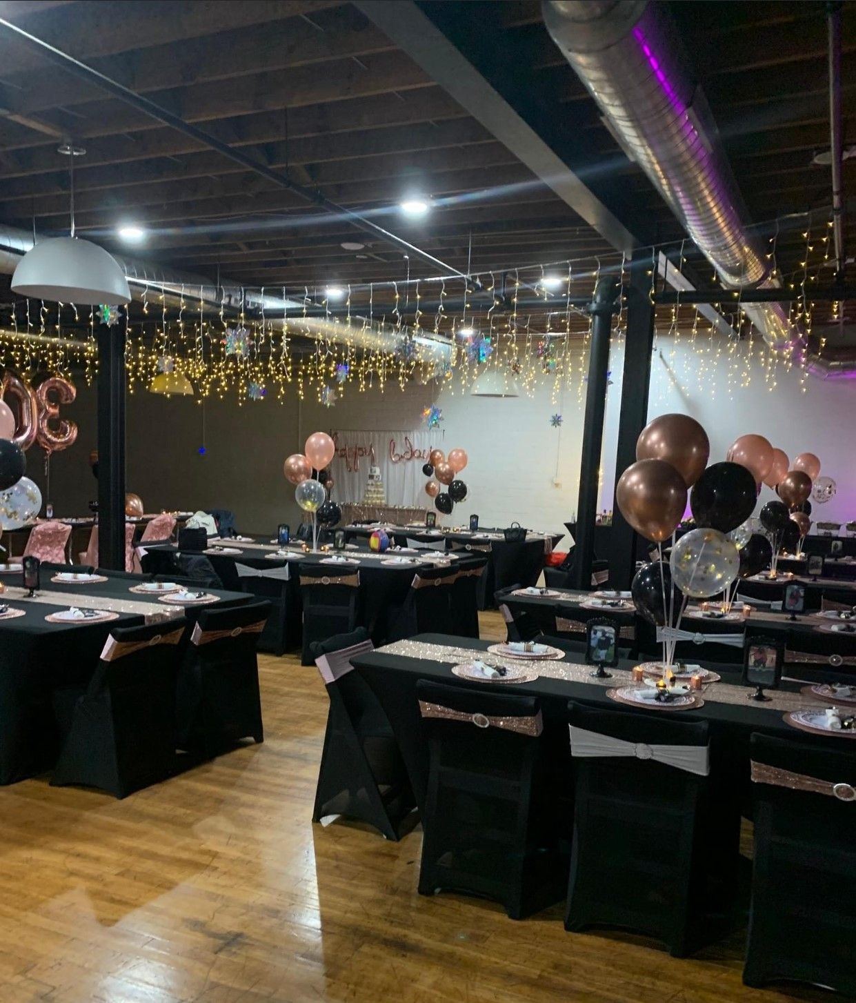 Event space decorated for a celebration with tables set, balloons, and string lights.