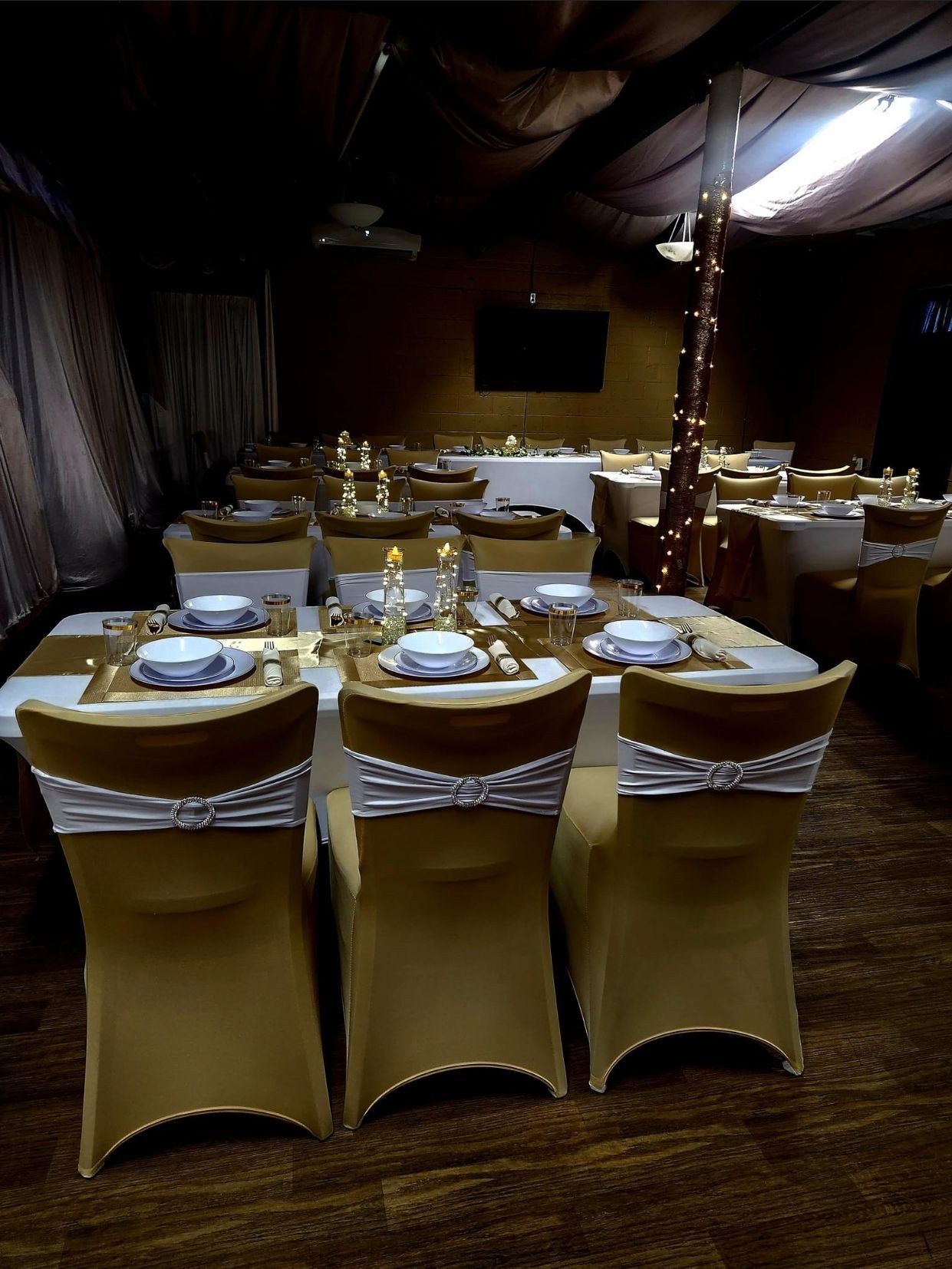 Gold-covered chairs around tables set for a formal event. String lights adorn a pole.